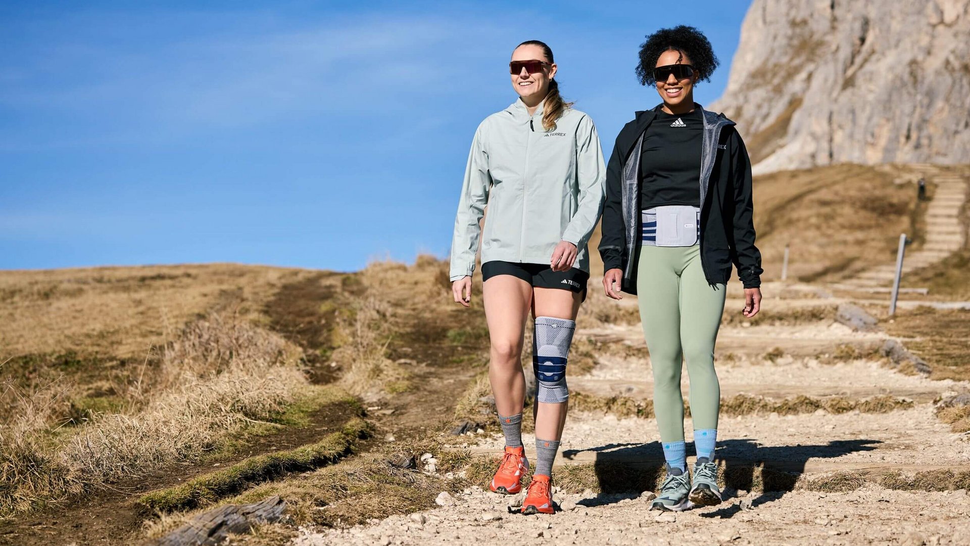 Laura Nolte und Deborah Levi wandern auf einem sonnigen Bergpfad mit felsigem Untergrund. Beide tragen Outdoor-Bekleidung und Sonnenbrillen; Laura trägt eine Kniebandage und Deborah eine Rückenbandage über ihrer Kleidung. Im Hintergrund sind ein trockener Grasbereich, ein steiler Pfad mit Stufen sowie felsige Bergformationen zu sehen.