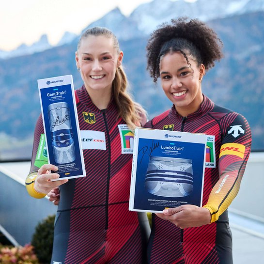 Laura Nolte und Deborah Levi, zwei Bob-Sportlerinnen in roten Rennanzuegen stehen im Freien vor einer Berglandschaft und halten Produktverpackungen von Bauerfeind (GenuTrain und LumboTrain) in die Kamera, beide laecheln.