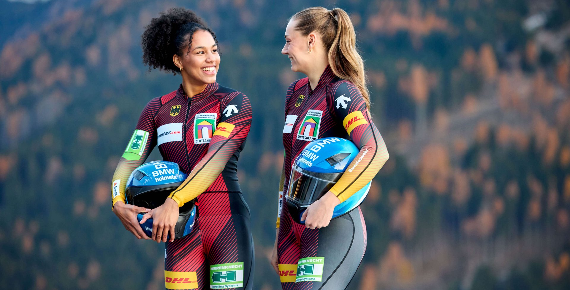Zwei Bobfahrerinnen, Laura Nolte und Deborah Levi, in roten Rennanzuegen stehen nebeneinander im Freien vor einer Bergkulisse und blicken in die Kamera, beide tragen Sportbekleidung mit Logos und wirken selbstbewusst.