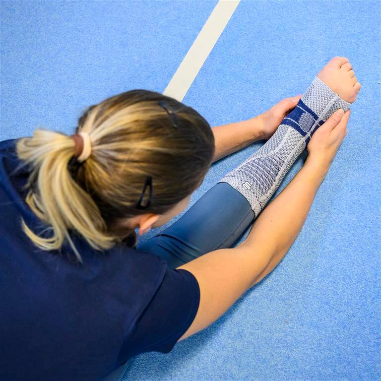 Turnerin dehnt ihr Bein auf einem blauen Boden und trägt die AchilloTrain-Bandage von Bauerfeind am rechten Fuß, die für die Unterstützung der Achillessehne entwickelt wurde.