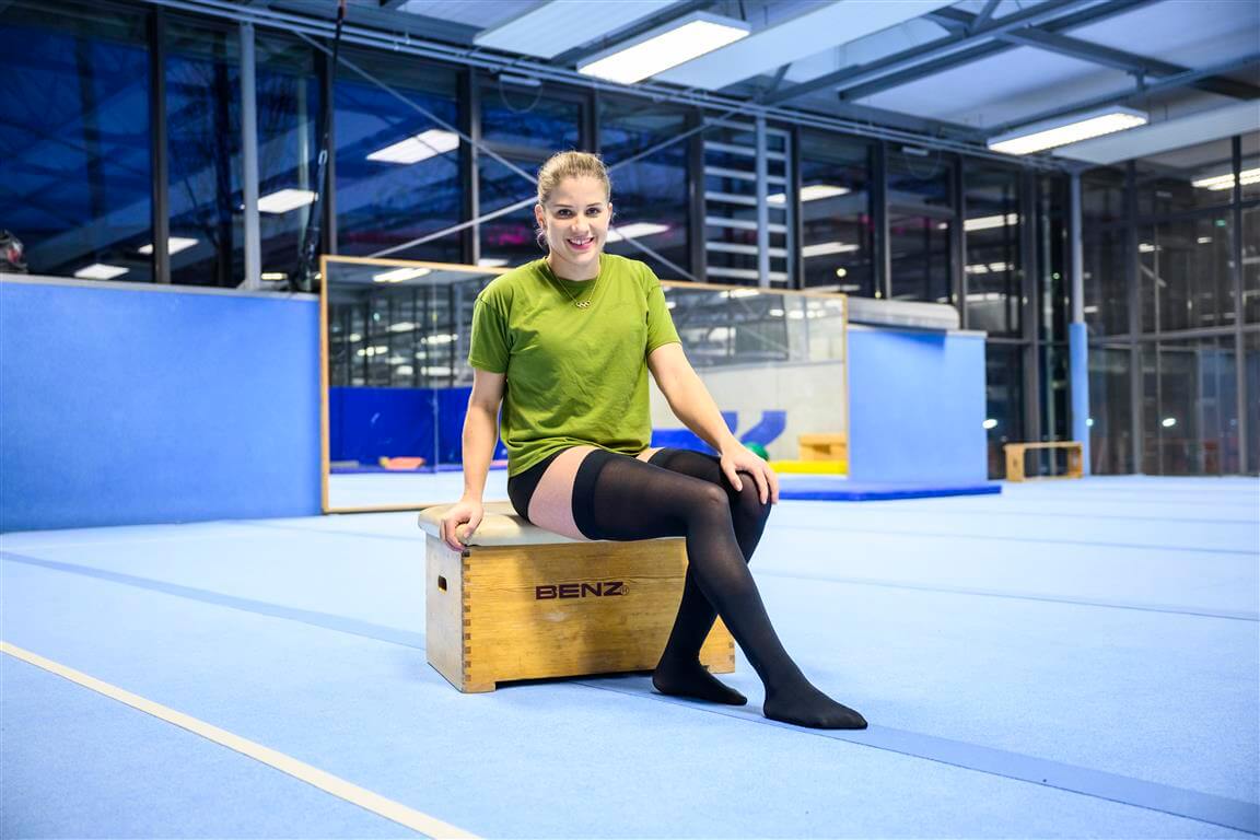Turnerin Elisabeth Seitz sitzt lächelnd auf einem Holzkasten in einer Sporthalle, trägt ein grünes T-Shirt und Bauerfeind Kompressionsstrümpfe, umgeben von blauer Trainingsausstattung.