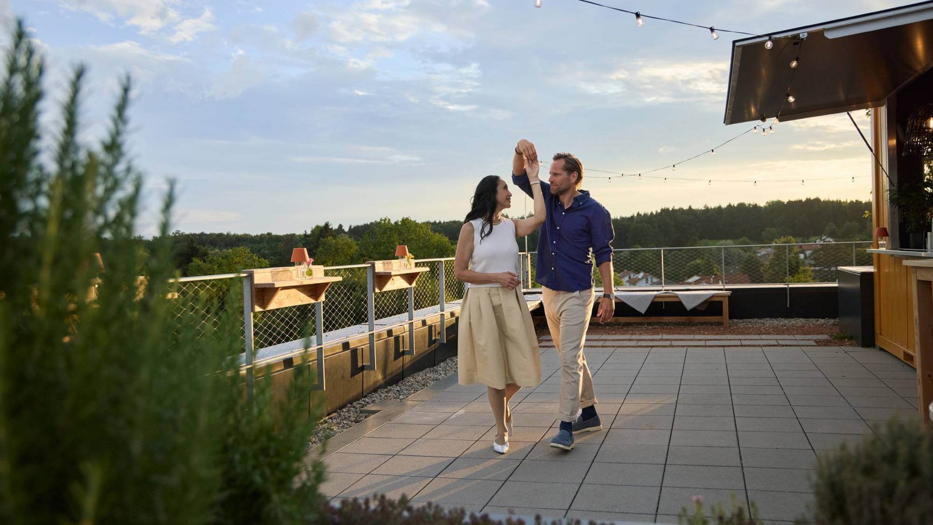 Paar tanzt auf einer Dachterrasse bei Abendstimmung unter Lichterketten mit Blick auf die umliegende Landschaft. Paar tanzt auf einer Dachterrasse bei Abendstimmung unter Lichterketten mit Blick auf die umliegende Landschaft.