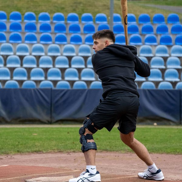 Simon Liedkte, Baseballprofi, steht in Schlagposition auf dem Baseballfeld. Er trägt die SecuTec Genu Flex Knieorthese, die sein verletztes Knie stabilisiert. Im Hintergrund sind leere, blaue Tribünensitze zu sehen. Simon konzentriert sich auf den bevorstehenden Schlag.
