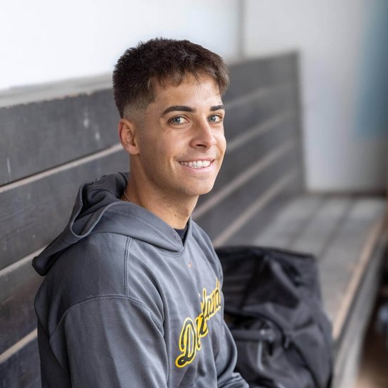 Baseballer Simon Liedkte sitzt lächelnd auf einer Holzbank im Dugout. Er trägt einen grauen Kapuzenpullover mit gelbem "Deutschland"-Schriftzug.