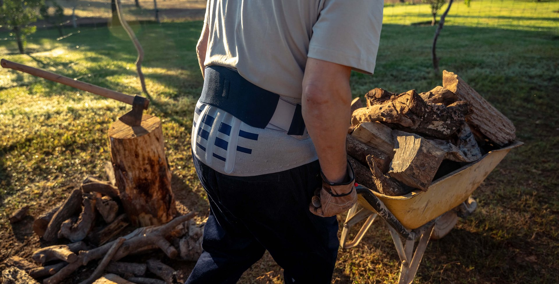 Ein Mann mit Rückenbandage von hinten, beim Bewegen einer gelben Schubkarre voller Holzstücke im Garten. Im Hintergrund steht ein Baumstumpf mit einer Axt, umgeben von Holzscheiten.
