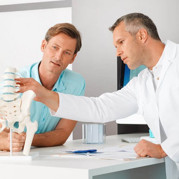Ein Arzt in weißem Kittel erklärt einem männlichen Patienten mithilfe eines Modells der menschlichen Wirbelsäule und des Beckens eine medizinische Diagnose. Der Patient hört aufmerksam zu und beide befinden sich in einem modernen Behandlungszimmer.