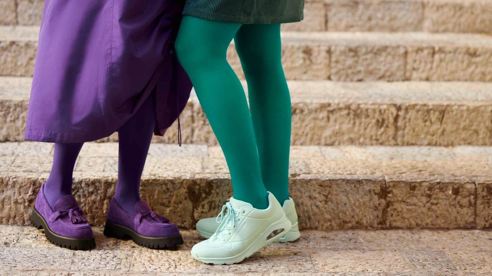 Nahaufnahme zweier Frauenbeine auf einer Steintreppe, betont durch auffällige VenoTrain Kompressionsstrümpfe in den Modefarben 'Irish Green' und 'French Lavender'.