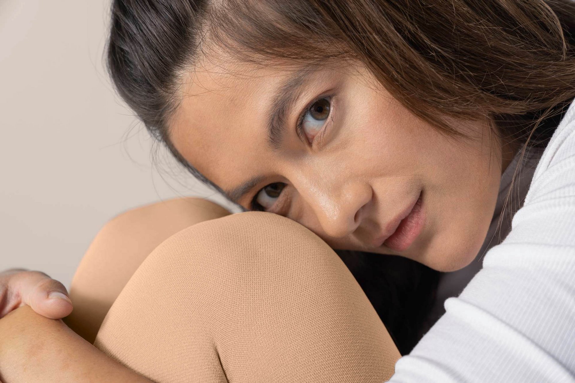 Nahaufnahme einer Frau mit beigen Strumpfhosen und weissem Oberteil, die ihren Kopf auf die angezogenen Knie legt und in die Kamera blickt. Nahaufnahme einer Frau mit beigen Strumpfhosen und weissem Oberteil, die ihren Kopf auf die angezogenen Knie legt und in die Kamera blickt.