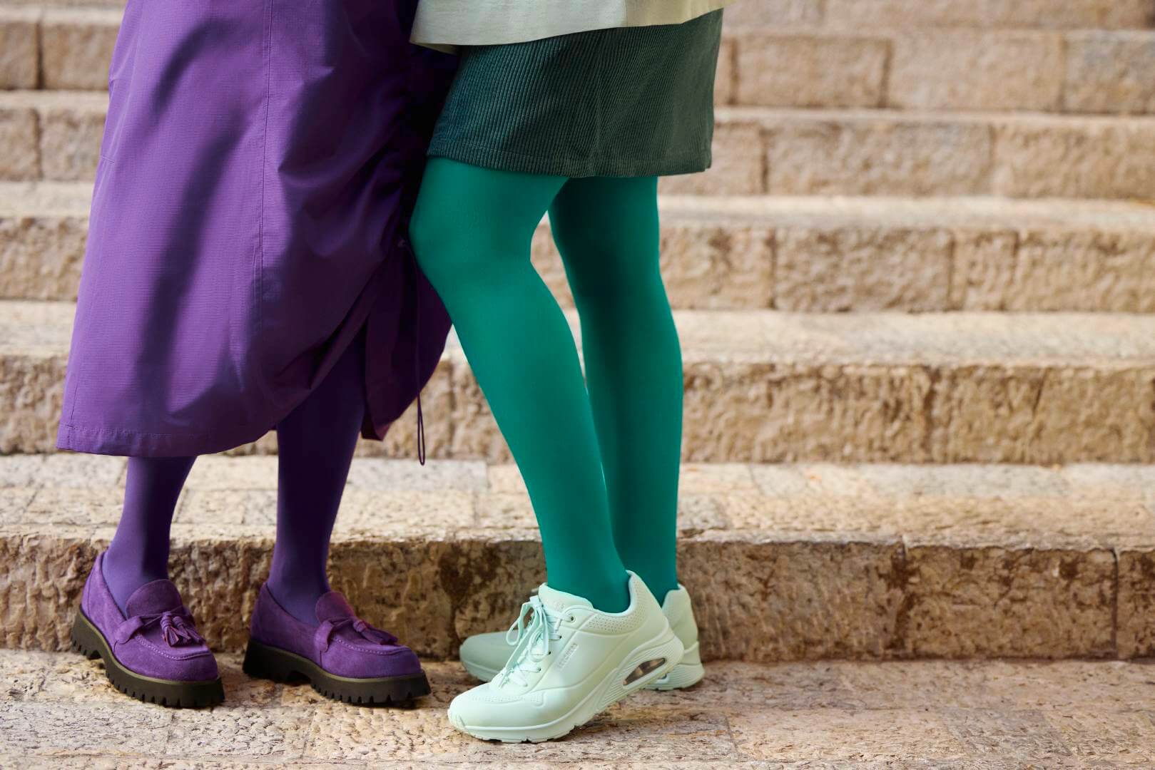 Nahaufnahme zweier Frauenbeine auf einer Steintreppe, betont durch auffällige VenoTrain Kompressionsstrümpfe in den Modefarben 'Irish Green' und 'French Lavender'.